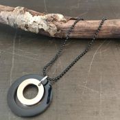 Sort og beige cirkulært halssmykke på sort kuglekæde, præsenteret på en rustik trægren.