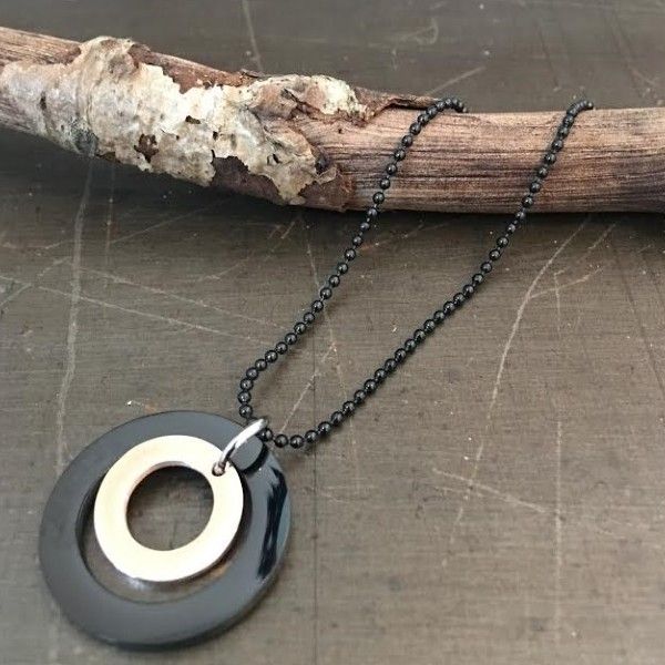 Sort og beige cirkulært halssmykke på sort kuglekæde, præsenteret på en rustik trægren.