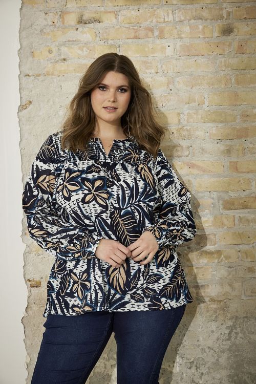 Elegant plussize bluse med bladprint og snøre i taljen, præsenteret foran en murstensvæg, perfekt til både hverdag og kontor.