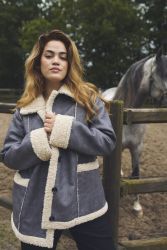 Elegant grå shearling jakke med imiteret pelskant, præsenteret udendørs med en hest i baggrunden.
