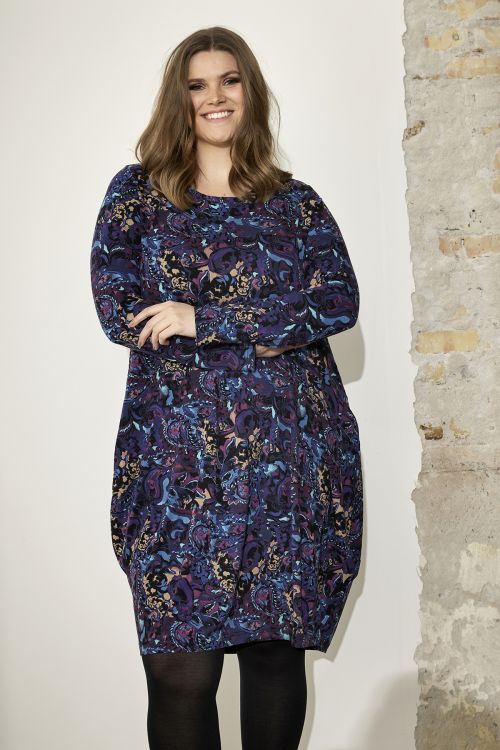 Plussize kjole med farverigt mønster, langærmet design og behagelig pasform, præsenteret foran en rå murstensvæg.