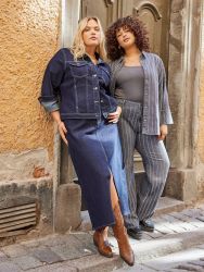 To modeller fremviser plussize mode med en denim nederdel og jakke samt et gråstribet sæt med blazer og bukser, stilfuldt koordineret med læderstøvler og sko.