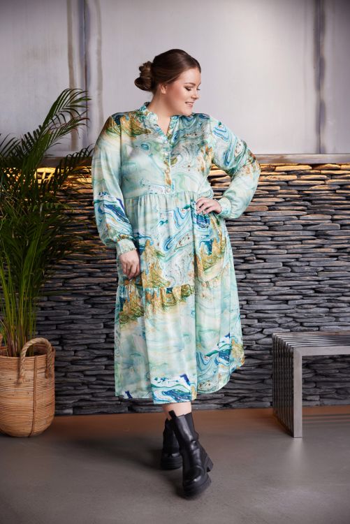 Plussize mønstret kjole med lange ærmer og krave, præsenteret i et moderne miljø med stenbaggrund og planter.