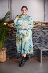 Plussize kjole med abstrakt mønster i blå og grønne nuancer, stylet med sorte støvler, poserer foran en stenbeklædt væg.