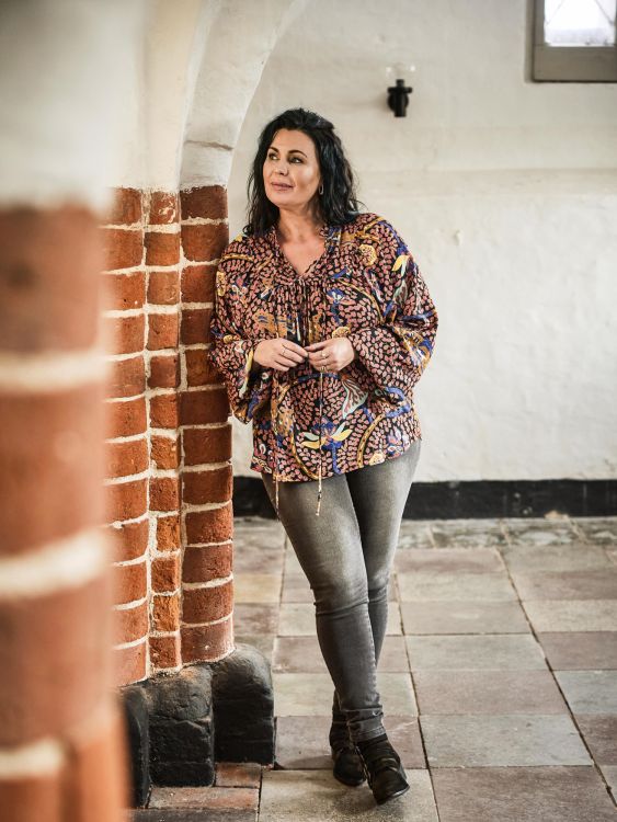 Plussize bluse med mønster i varme farver kombineret med grå skinny jeans og støvler, præsenteret i en rustik indstilling.