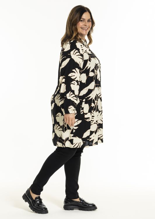 Plussize blomstret tunika i sort og beige med lange ærmer og rund hals, kombineret med sorte, kropsnære bukser og sorte snøresko.