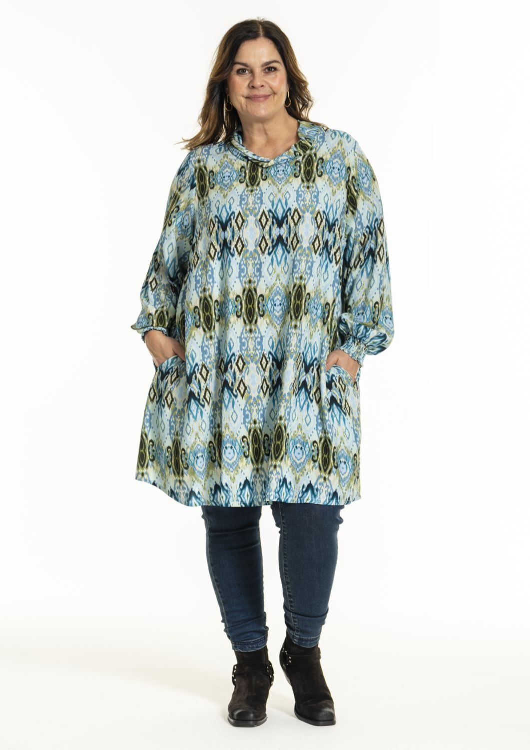 Plussize tunika med etnisk mønster og trekvartærmer, kombineret med mørke slim jeans og sorte støvler.