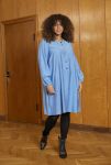 Blå oversize skjortekjole med broderi detaljer kombineret med sorte leggings og spidse støvler på trægulv mod trædør baggrund.