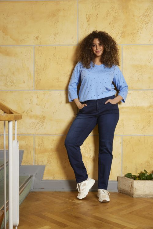 Moderigtigt plussize outfit med prikket bluse og marineblå jeans, præsenteret i et indendørs miljø med trægulv og gul væg.