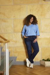 Plussize outfit præsenteret af model foran en gul væg: blå prikket bluse og mørkeblå bukser med hvide sneakers. Stilfuld og afslappet mode.